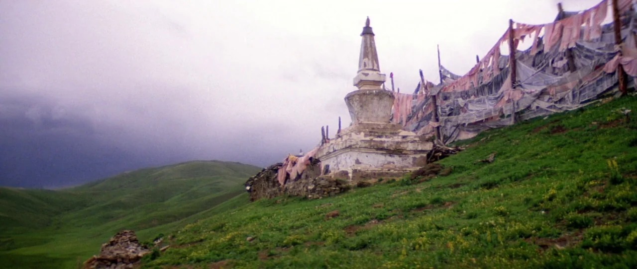 Horse Thief movie still: wide — A weathered stone stupa sits on a grassy mountainside under a heavy, overcast sky. Rows of frayed, faded prayer…