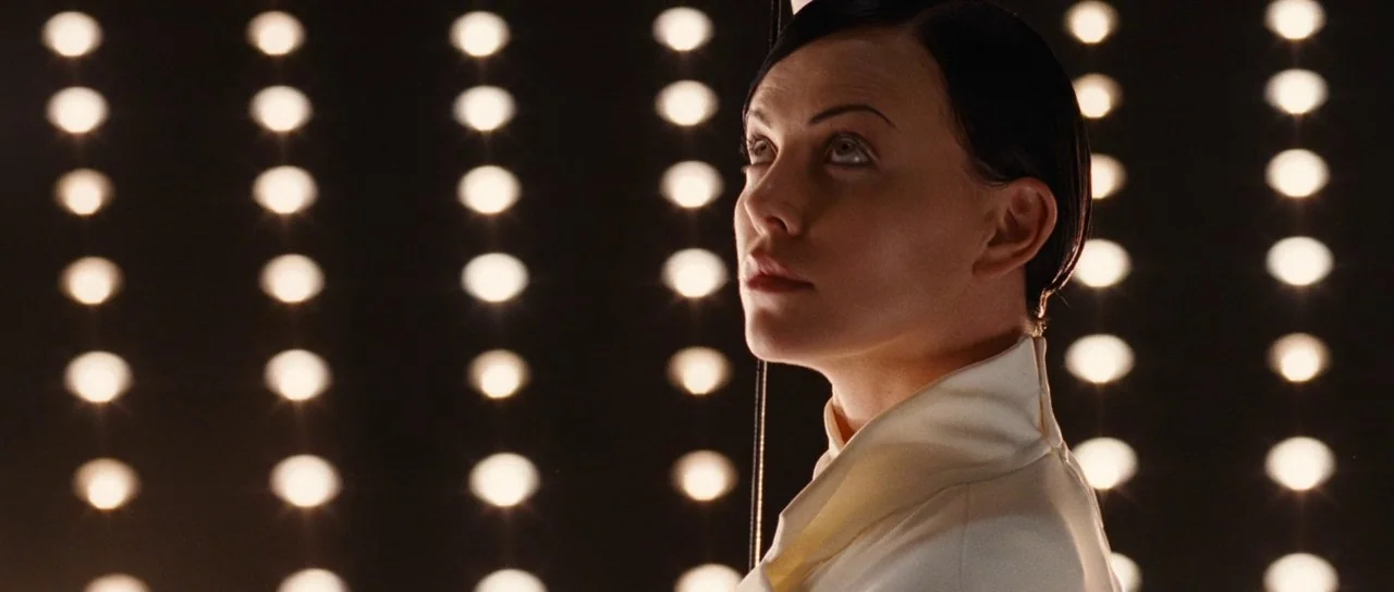Aeon Flux (2005) movie still: closeup — The subject stands in front of a dark wall filled with rows of lights. The woman looks upward with a direct…