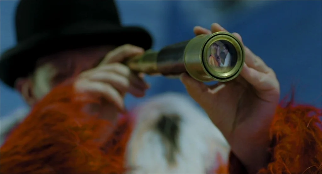 The Fall movie still: closeup — A figure wears a bowler hat and a shaggy orange coat in a studio. They peer through a brass telescope with a small…