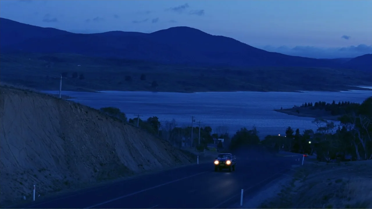 Emily (1976) movie still: wide — A rural highway cuts through hilly terrain bordered by a lake under blue twilight. A single pickup truck drives…