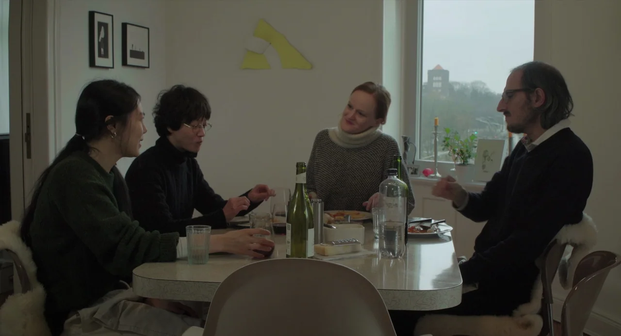 On The Beach At Night Alone (2017) movie still: medium — A light-colored dining room features a window overlooking a cityscape in the distance. Four…