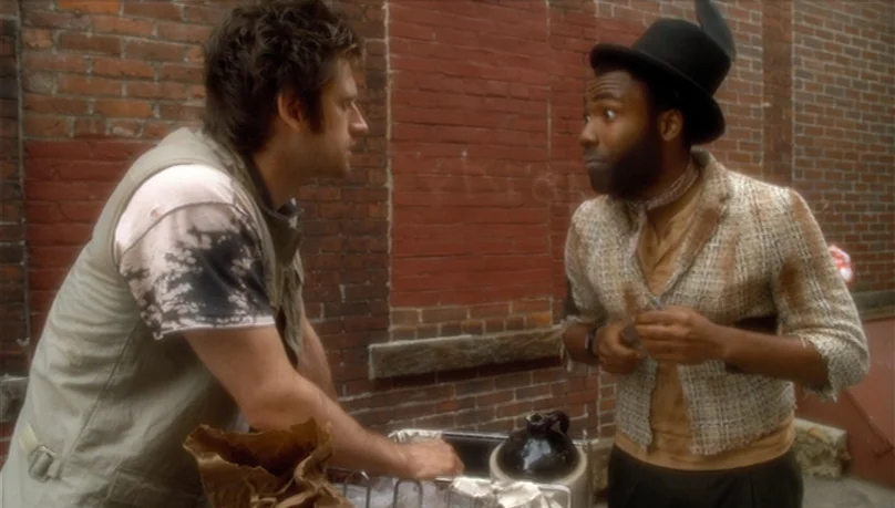 Mystery Team (2009) movie still: medium — Two men stand together against a weathered red brick wall in a paved area. One man wears a top hat while…