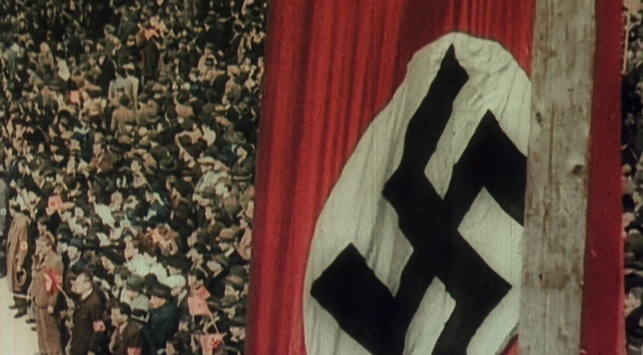 Underground (1995) movie still: wide — A massive crowd of people fills a stadium during a public gathering. A large red banner featuring a black…