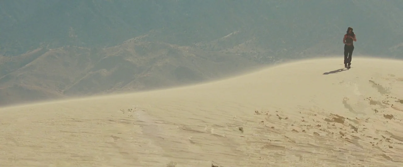 Iron Man (2008) movie still: wide — A wide desert landscape features massive undulating sand dunes under a clear, bright sky. A solitary man walks…