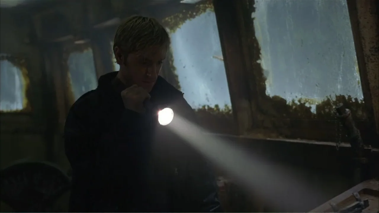 Ghost Ship (2002) movie still: medium — A dark, decaying boat cabin shows signs of water damage and peeling paint. A young man directs a beam of…