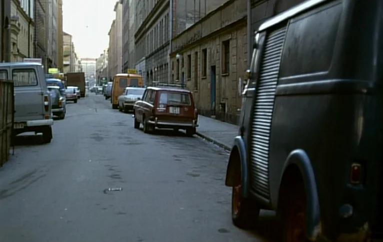 Giliap (1975) movie still: wide — A narrow cobblestone street lined with multi-story apartment buildings and parked vintage vehicles. Several cars…