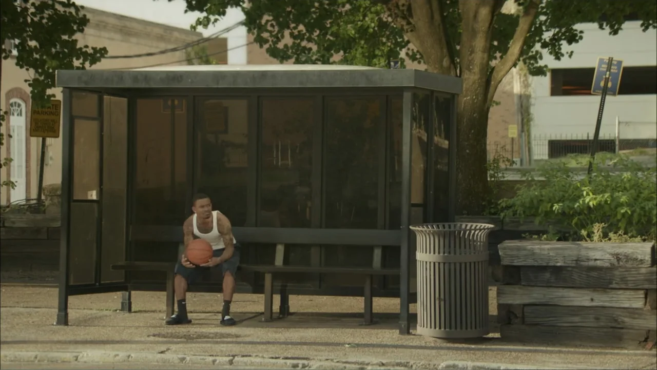 The Hollars (2016) movie still: medium — A metal and glass bus shelter sits on a concrete sidewalk beside a tree. A man in a tank top sits on the…