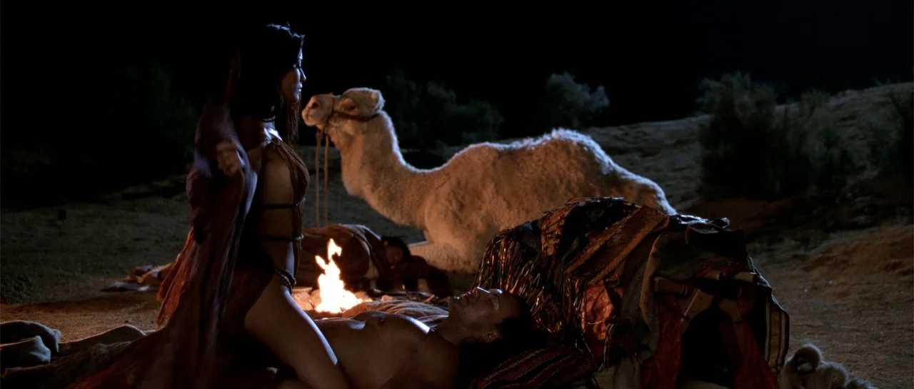The Scorpion King (2002) movie still: medium — An open desert terrain features a small campfire flickering beside two resting people. A woman kneels…