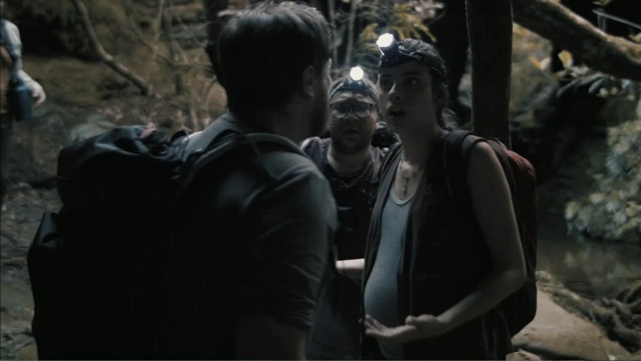 The Grotto (2022) movie still: medium — A damp, dark, and uneven ravine area serves as the setting for three people hiking. A pregnant woman stands…