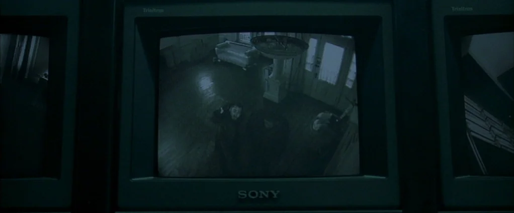Panic Room (2002) movie still: wide — A high-angle security camera view displays a dark residential entryway with a sofa present. Two men stand in…