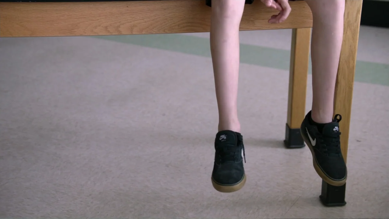 Judy Blume Forever (2023) movie still: medium — A wooden table sits on a linoleum floor in a well-lit indoor space. A child sits on the edge of the…