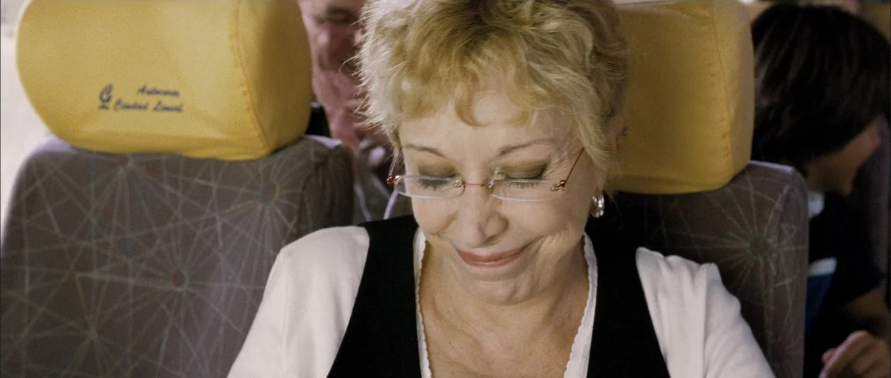 Rivals (1972) movie still: medium — An interior of a passenger bus with yellow headrest covers and patterned upholstery. An elderly woman sits in a…