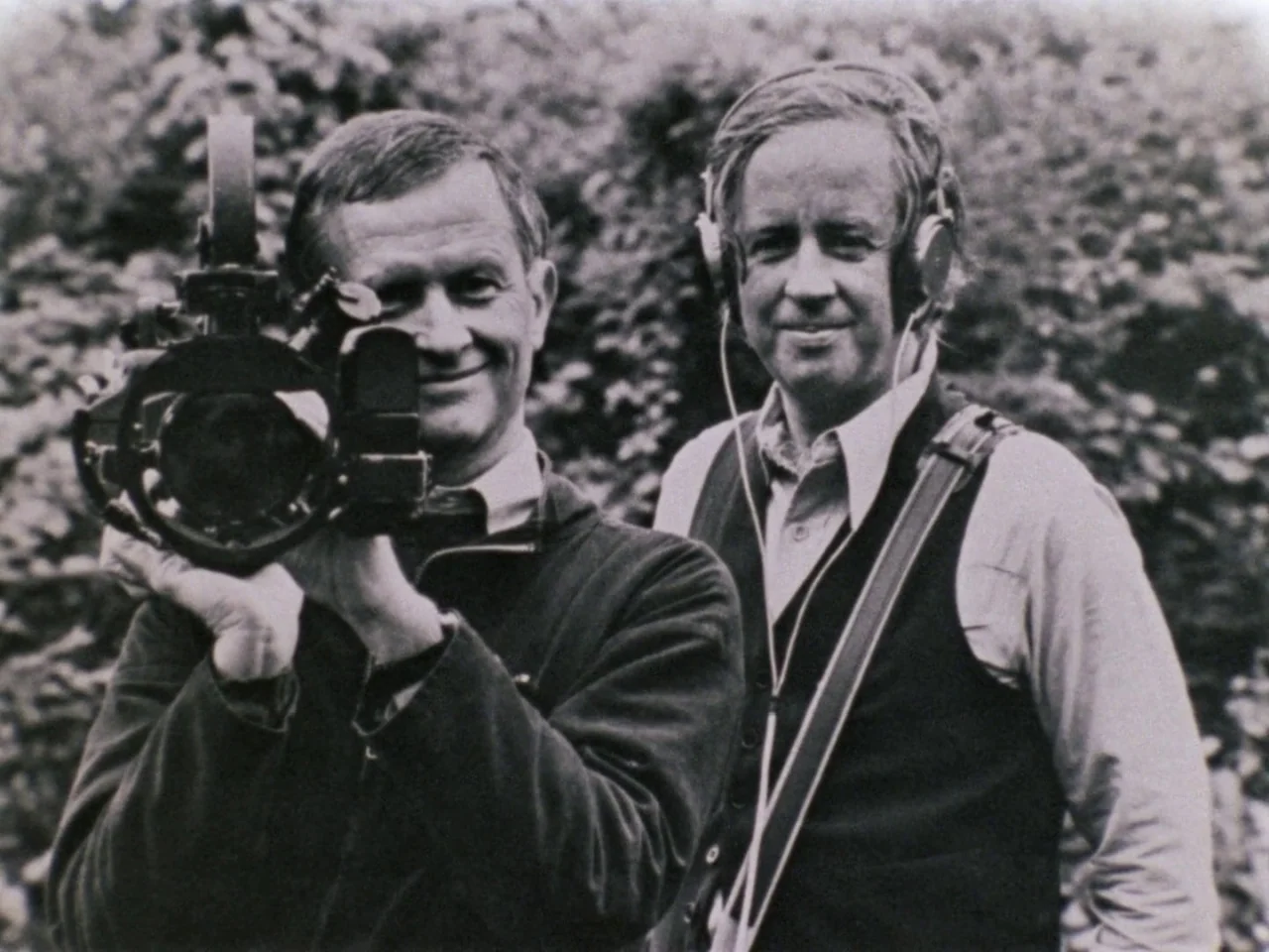 Grey Gardens (1975) movie still: medium — Two men stand outdoors before a dense wall of leafy foliage. One man holds a movie camera while the other…