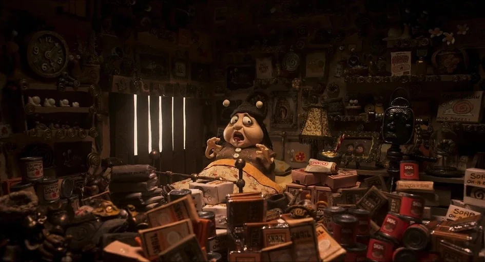 Memoir of a Snail (2024) movie still: medium — A cluttered, cramped room is packed with stacks of tins and boxes everywhere. A woman sits upright in…