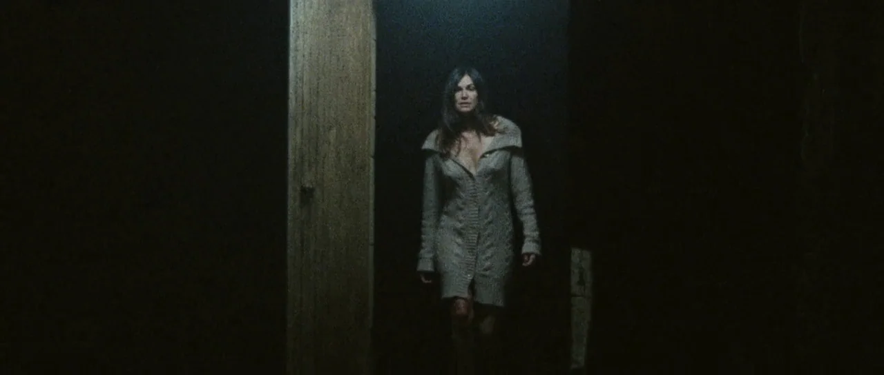 Alléluia (2014) movie still: medium — A woman stands in a dimly lit, narrow corridor with dark walls. She wears a light-colored knit sweater dress…