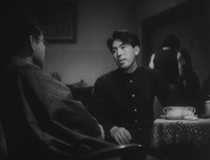 No Regrets for Our Youth (1946) movie still: medium — A dark dining room with a lacy tablecloth and a ceramic bowl on top. Two men sit at the table…