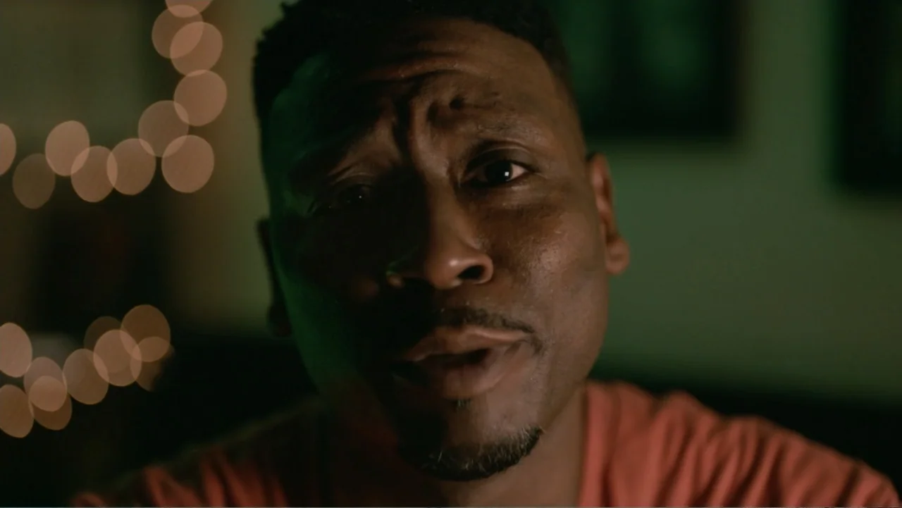 Between the World and Me (2020) movie still: closeup — A dark interior space features a man in an orange shirt. He stands directly in front of the…