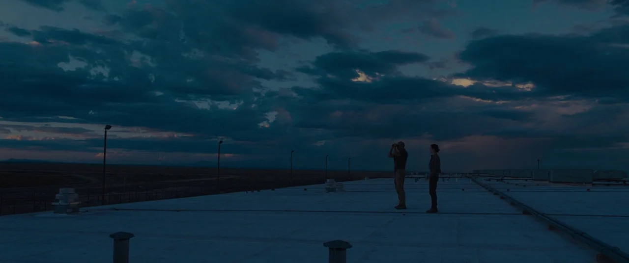 Sicario (2015) movie still: wide — A flat gravel rooftop overlooks an expansive desert landscape under a dark cloudy sky. A man using binoculars…