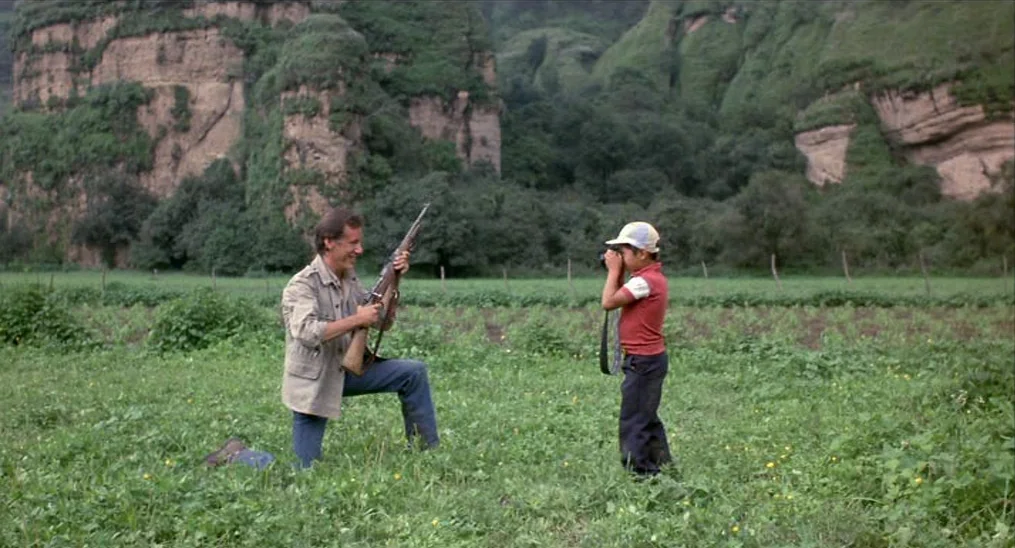 Salvador (1986) movie still: fullbody — A green pasture lies beneath high, rugged rock cliffs in a rural landscape. A kneeling man holds a rifle…