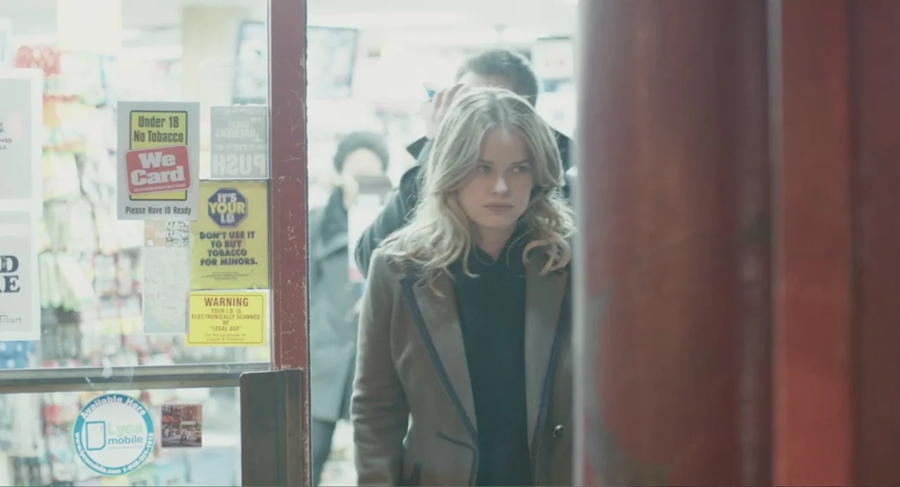 Before We Go (2014) movie still: medium — A glass door covered in informational signs reveals the interior of a brightly lit shop. A woman stands in…