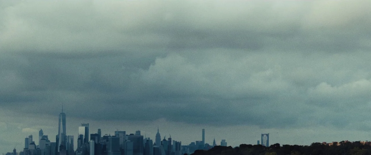 Daniel Isn’t Real movie still: establishing — A low-angle view of the New York City skyline under heavy cloud cover. Numerous skyscrapers…