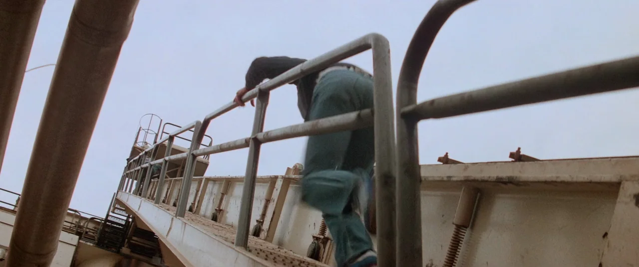 Vigilante (1982) movie still: fullbody — An industrial metal catwalk extends upward against a pale gray sky. A person climbs the ladder while…