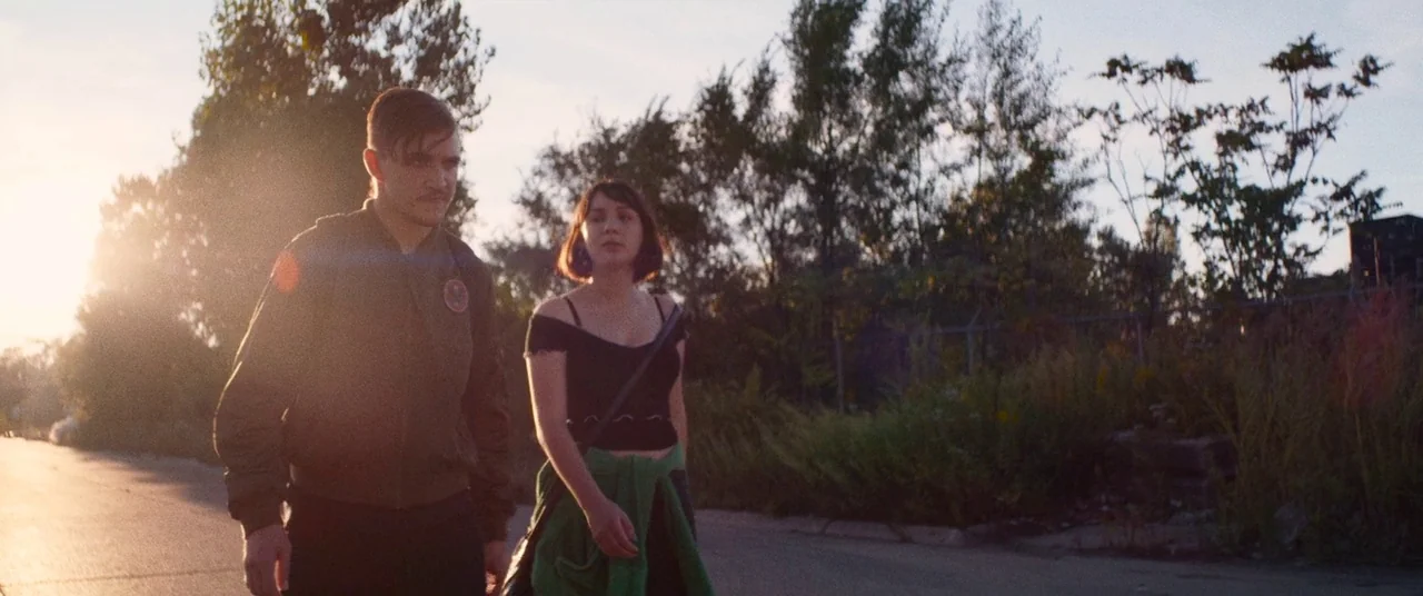 Dinner in America (2020) movie still: fullbody — A paved road bordered by overgrown grass and trees during a bright sunset. A man and a woman walk…