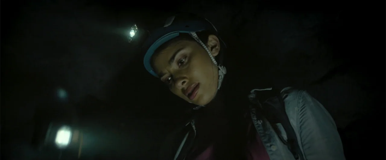 Black Water: Abyss (2020) movie still: closeup — A dark, rocky cave interior is illuminated only by a personal headlamp. A woman wearing a helmet…