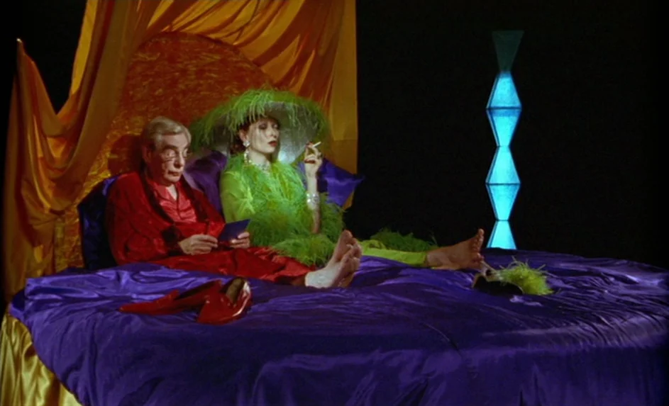 Wittgenstein (1993) movie still: medium — A dark, theatrical bedroom features a large bed with deep purple satin sheets. The man and woman sit…