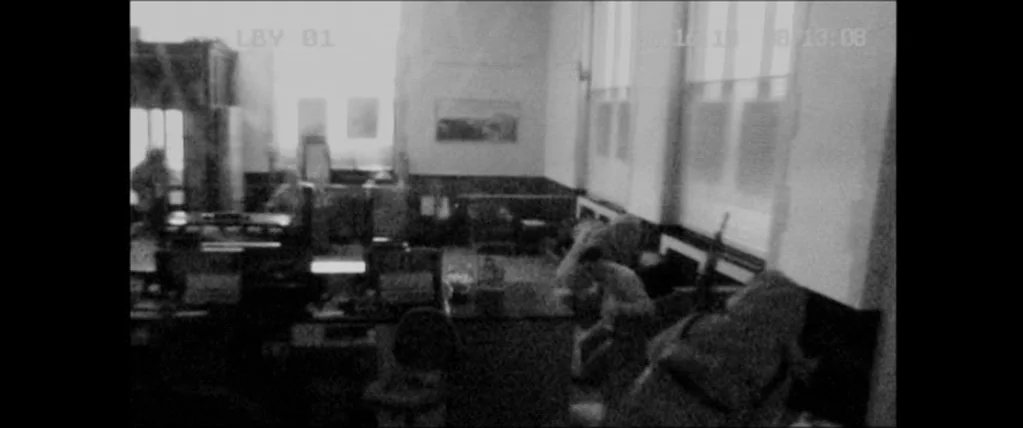 The Town (2010) movie still: wide — A low-resolution view of a commercial bank floor featuring desks and tall windows. The space appears empty with…
