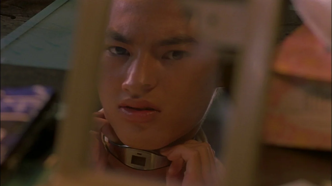 Battle Royale (2000) movie still: closeup — A teenage boy stares into a mirror while adjusting an electronic collar around his neck. The reflection…
