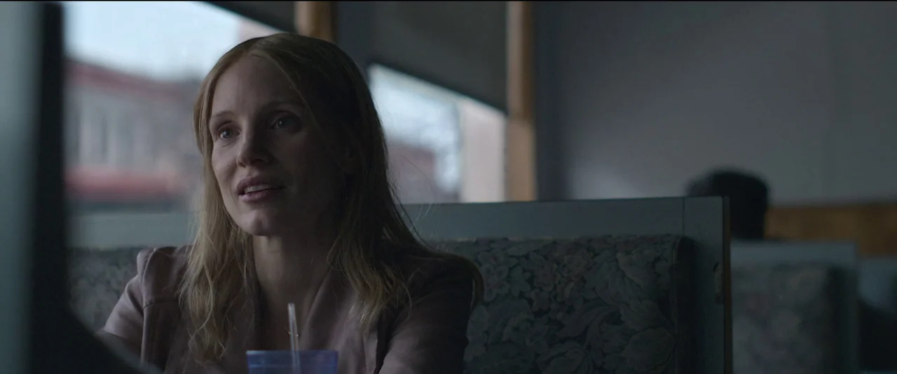 The Good Nurse (2022) movie still: medium — A woman sits inside a booth at a dim diner with windows in background. She is looking forward while…