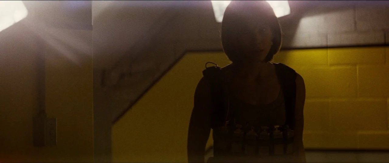 Speed (1994) movie still: medium — A dimly lit stairwell with yellow painted cinder block walls and industrial architectural features. A woman in a…