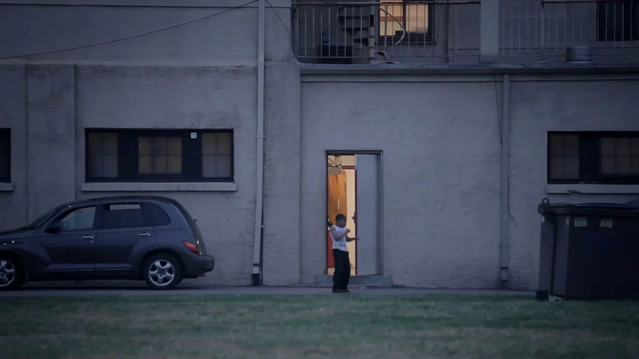 T-Rex (2015) movie still: wide — The exterior of a beige building at dusk features a car and a dumpster. A teenager stands in the open doorway of…