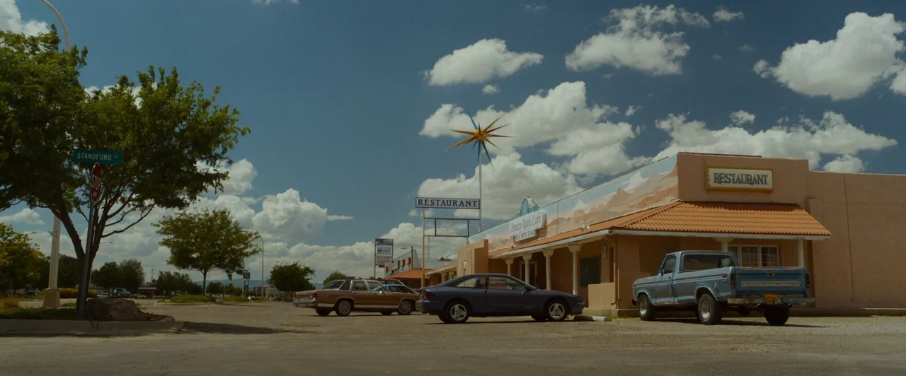 The Guest (2014) movie still: wide — A suburban parking lot featuring a low-rise tan stucco restaurant with a mural. Three parked vehicles sit in…