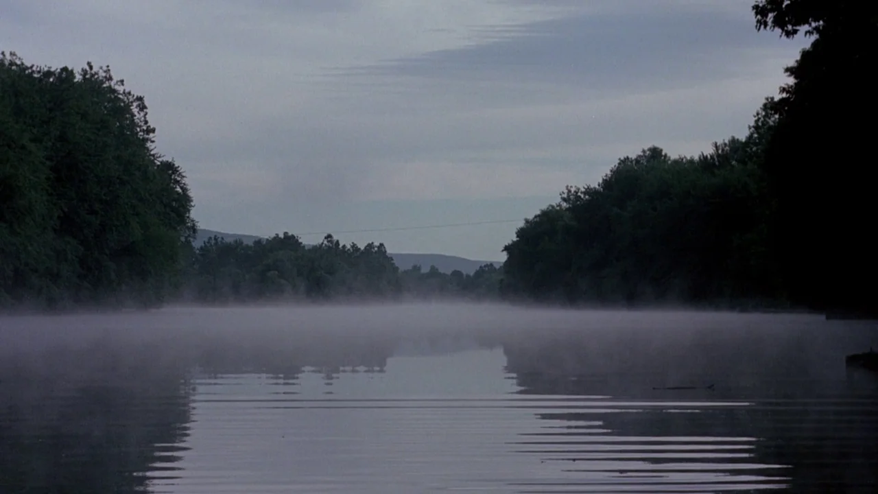 I Spit On Your Grave (1978) movie still: establishing — A wide, still river flows between two dense, tree-lined banks under an overcast sky. A thin…