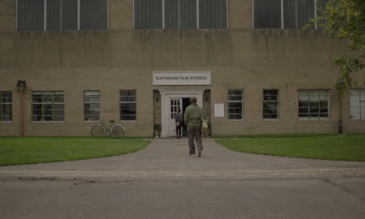 The Souvenir (2019) movie still: wide — A stone institutional building has a sign reading Raynham Film School above the door. A person in an olive…