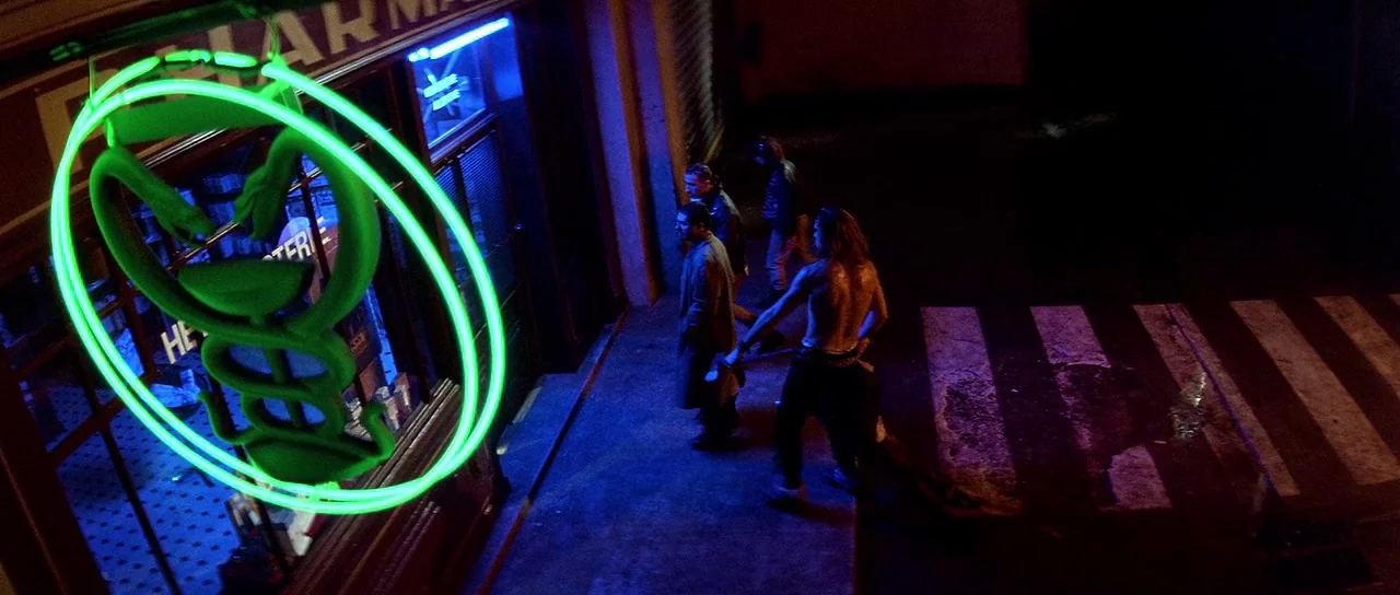 Nikita (1990) movie still: wide — A city sidewalk at night is illuminated by a prominent glowing green neon caduceus sign. Three men walk past the…