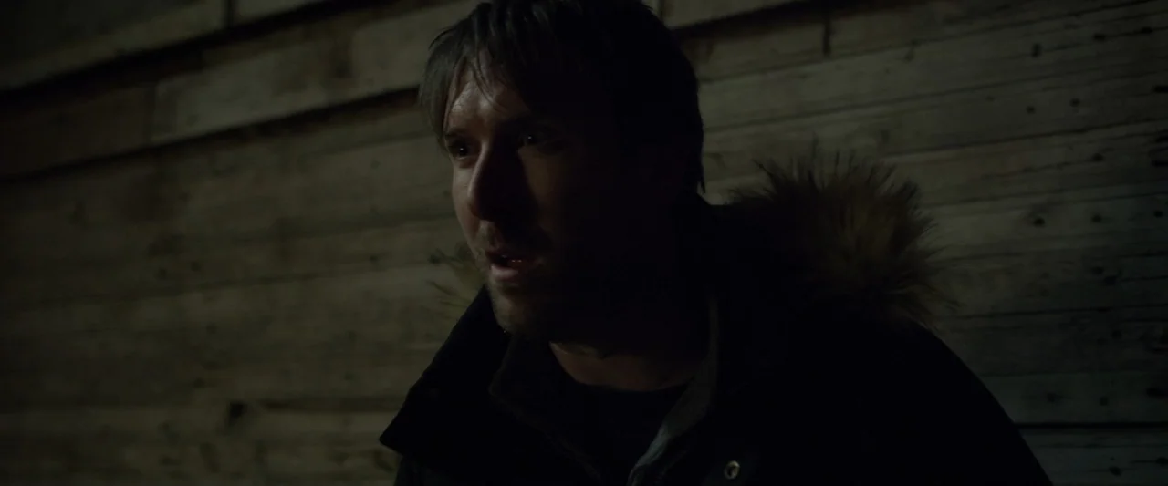 Motherly (2022) movie still: medium — A dark, cramped interior space features horizontal wooden plank walls. A man in a dark winter coat looks…