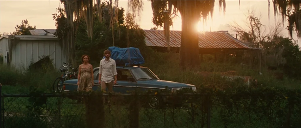 Away We Go (2009) movie still: wide — An overgrown yard with a blue car, a small shed, and moss-draped trees. A pregnant woman and a man walk toward…