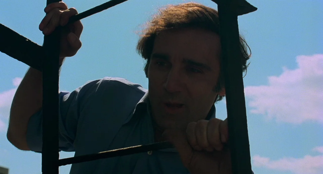 God Told Me To (1976) movie still: medium — A man climbs a metal ladder against a bright blue sky background. He grips the iron rungs while looking…