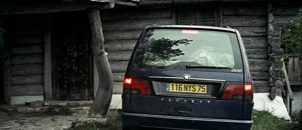 Time Of The Wolf (2003) movie still: wide — A dark blue Peugeot minivan is parked in front of a rustic log cabin. The vehicle is positioned on a…