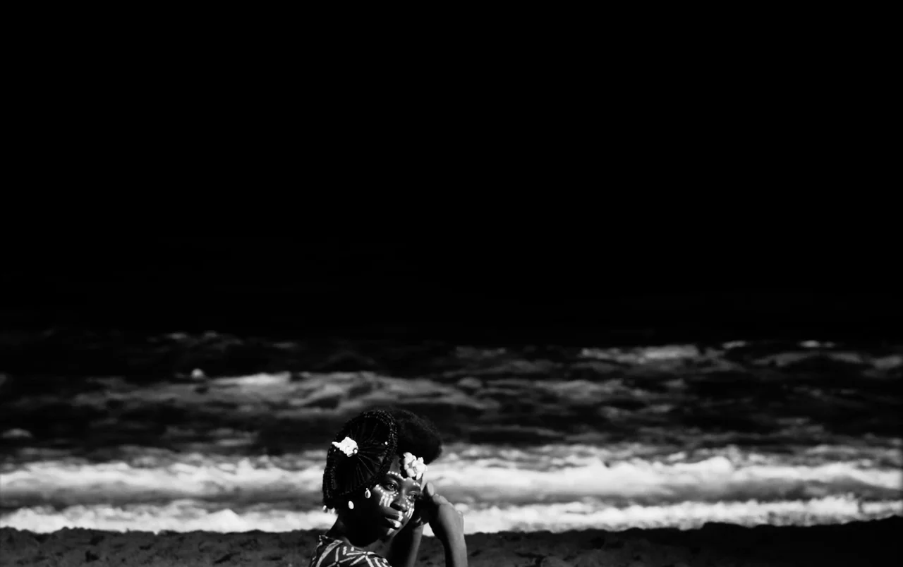 Mami Wata (2023) movie still: medium — A dark sandy coastline meets turbulent ocean waves during a high-contrast daylight session. A woman with…