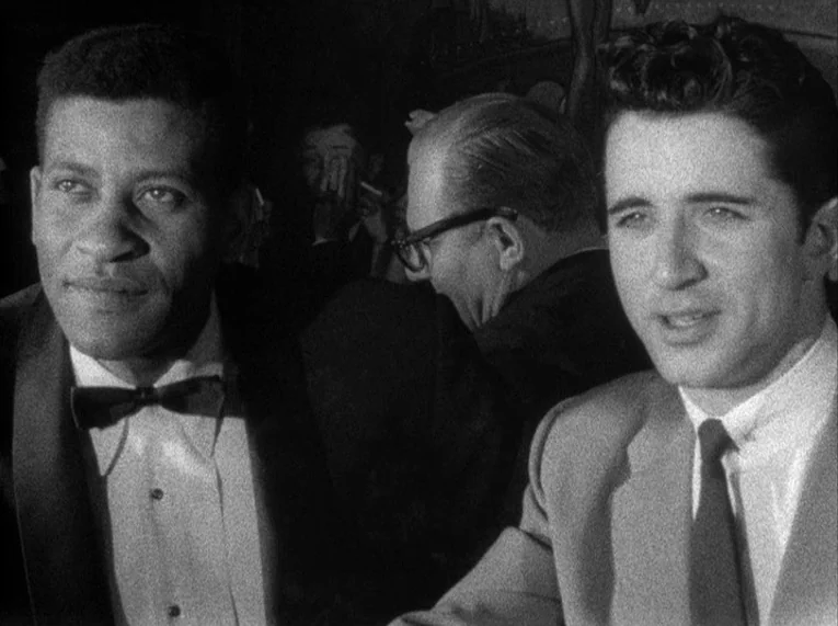 Shadows (1958) movie still: medium — A dimly lit club with guests seated at tables in the dark background. Two men in formal attire sit in the…