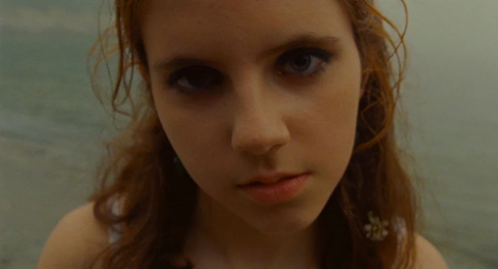 Moonrise Kingdom (2012) movie still: closeup — A teenage girl stands outdoors near a body of water in muted natural light. She looks directly at the…