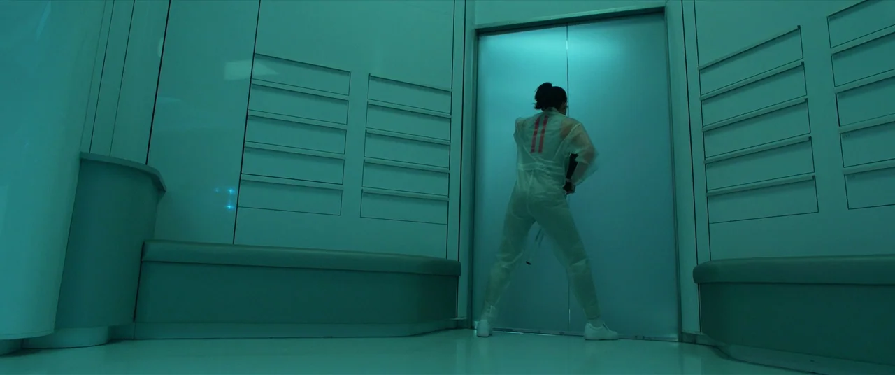 The Predator (2018) movie still: fullbody — A clinical, futuristic corridor features symmetrical wall panels and a central sliding door. A person in…