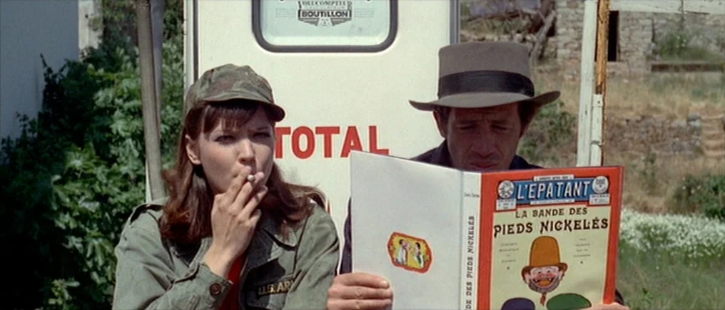 Pierrot Le Fou (Crazy Pete) (1965) movie still: medium — A woman and a man sit in front of a vintage gasoline pump outdoors. The woman smokes a…