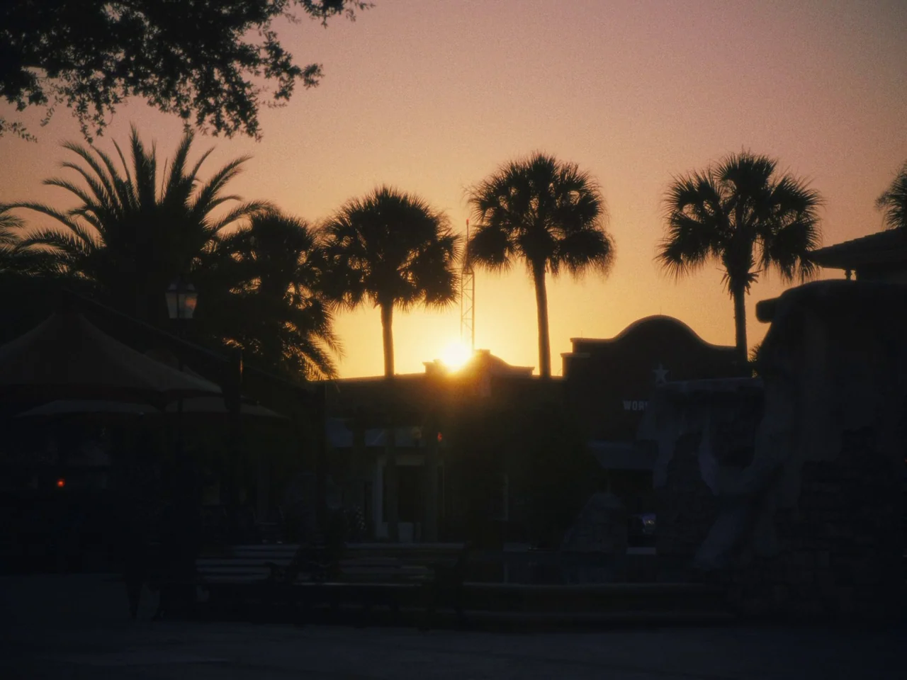 Some Kind Of Heaven (2020) movie still: wide — An outdoor plaza features silhouettes of palm trees and low buildings against a sunset. The sun sits…