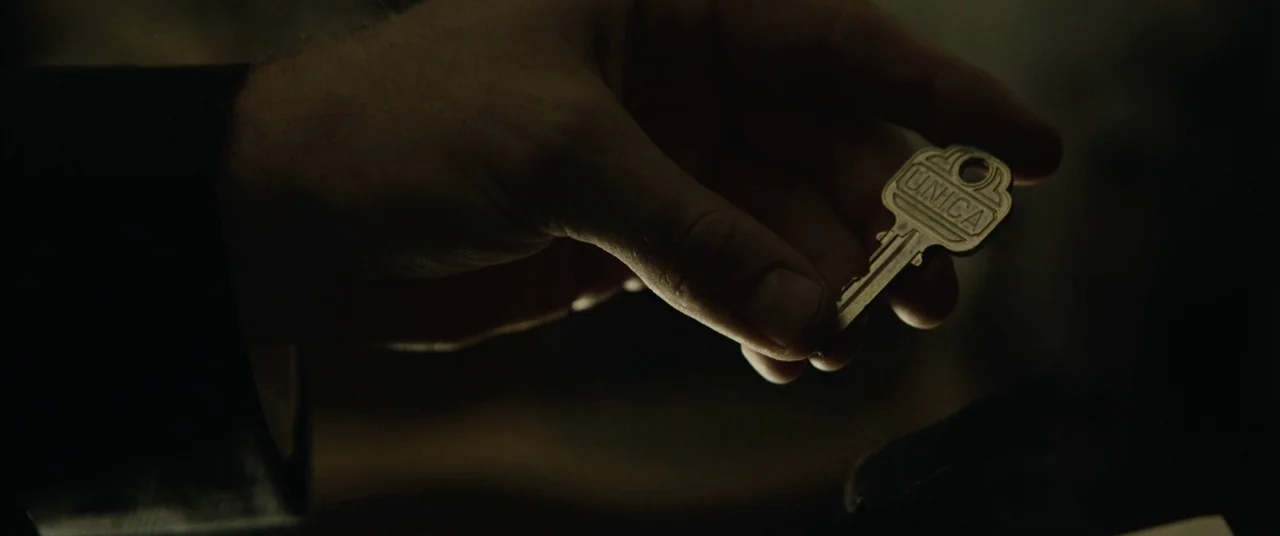 Enemy (2013) movie still: closeup — A dimly lit interior space features a hand extending toward the foreground. The fingers hold a silver key with…