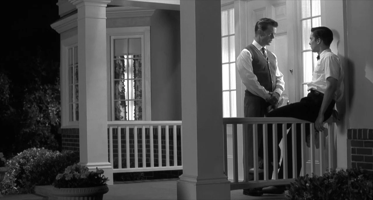 Pleasantville (1998) movie still: fullbody — A suburban house porch at night with brick foundations and white wooden railings. Two men stand…